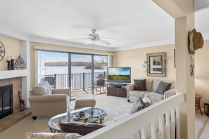A spacious living room with a large TV, comfortable seating, a fireplace, and a balcony with a view of a lake.