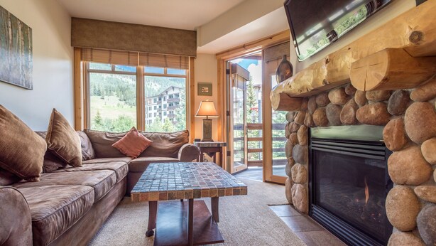 A cozy living room with a stone fireplace, a leather sofa, and a view of a mountainous landscape through large windows. - Beautiful one bedroom condo in the heart of the center village. (Copper Mountain)