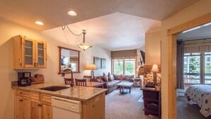 A spacious kitchen with wooden cabinets, a granite countertop, and a dishwasher. There is a dining area with a table and chairs, and a living room with a sofa and a fireplace. - Beautiful one bedroom condo in the heart of the center village. (Copper Mountain)