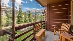 A balcony with a wooden bench and chairs, offering a view of a mountainous landscape with trees and buildings. - Beautiful one bedroom condo in the heart of the center village. (Copper Mountain)