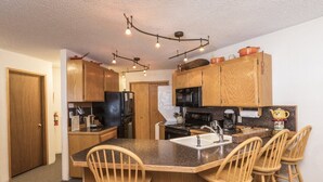 Une cuisine compacte avec des meubles en bois, un réfrigérateur noir, un four à micro-ondes et une cuisinière. On y trouve quatre tabourets de bar en bois disposés autour d’un îlot central doté d’un évier.