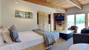Ein Schlafzimmer mit Holzboden, einem Bett mit Kissen, einem Fernseher und Blick ins Freie durch große Fenster.