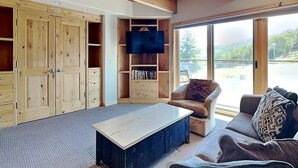 Ein gemütliches Wohnzimmer mit Holzbalkendecke, einem Flachbildfernseher, einer Couch und einem großen Fenster mit Blick ins Freie.