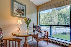 A room with a round glass table, a chair, a lamp, a vase with flowers, and a framed picture on the wall.