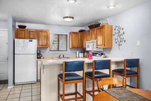 A compact kitchen with wooden cabinets, a white refrigerator, a microwave, and a sink. There are three blue chairs around a small wooden table.