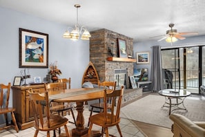 A dining area with a stone fireplace, wooden chairs, a chandelier, and a television.