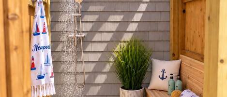 A wooden pergola with a window, a showerhead, a potted plant, a bench with pillows, and a rug on a tiled floor.