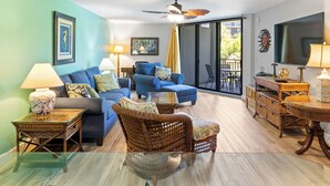 A living room with a sofa, two armchairs, a glass coffee table, wicker side tables, a ceiling fan, and a TV.