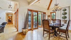A spacious dining area with a wooden table and chairs, a chandelier, and a view of the outdoors through large glass doors. - La Vita Dolce – Romantic Carmel-by-the-Sea Cottage Near Beach & Ocean Ave (Carmel by the Sea)