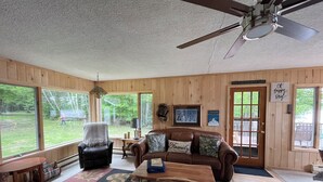 A cozy room with a wooden interior, a ceiling fan, a couch with a family pillow, a table, and a large window overlooking a yard.