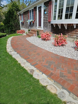 A house with a brick pathway leading to the front door, surrounded by a well-maintained lawn and flowering shrubs.