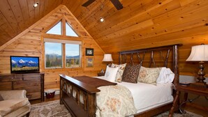 Ein Schlafzimmer mit Holzboden, einem großen Bett mit vielen Kissen, einem Fernseher auf einer Kommode und einem Fenster mit Blick auf die Berge.