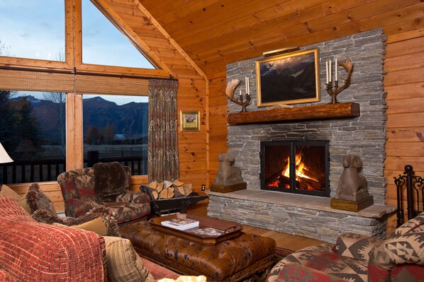 A cozy log cabin interior with a stone fireplace, wooden furniture, and a scenic mountain view through large windows.