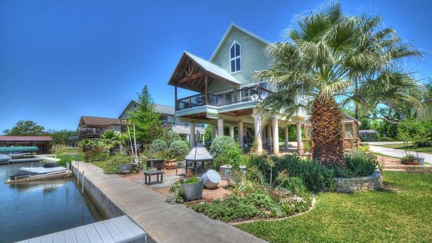 A large house with a covered porch, a swimming pool, and a well-maintained garden with palm trees and various plants. - Monthly rental getaway on lake LBJ (Kingsland)