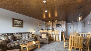 Una sala de estar acogedora con un sofá, sillones y una mesa de centro. Se observa una zona de cocina con armarios de madera y un microondas.