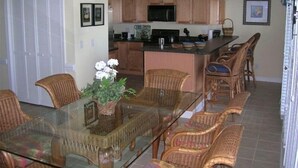 A dining area with a glass-top table and wicker chairs, a kitchen with wooden cabinets, and a small island with a microwave. -  Beautiful 4 BR Oib Condo with Beachfront Pool!  (Ocean Isle Beach)