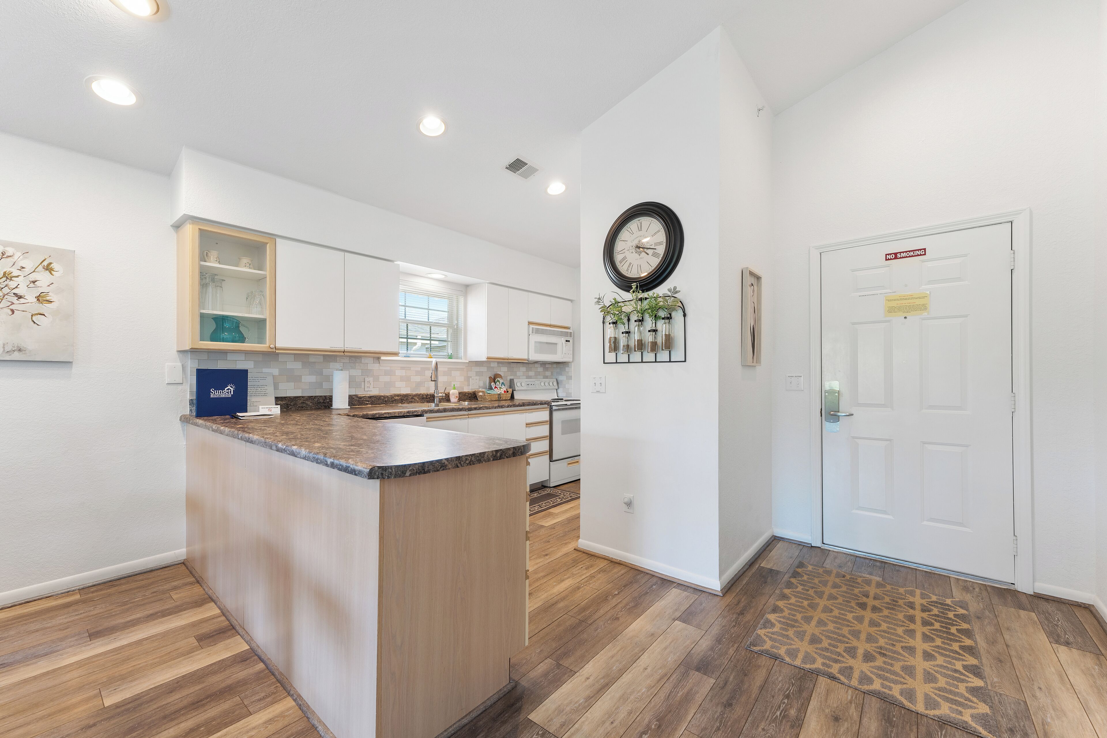 A modern kitchen with a central island, white cabinetry, and a patterned rug leading to a door with a 'No Smoking' sign.