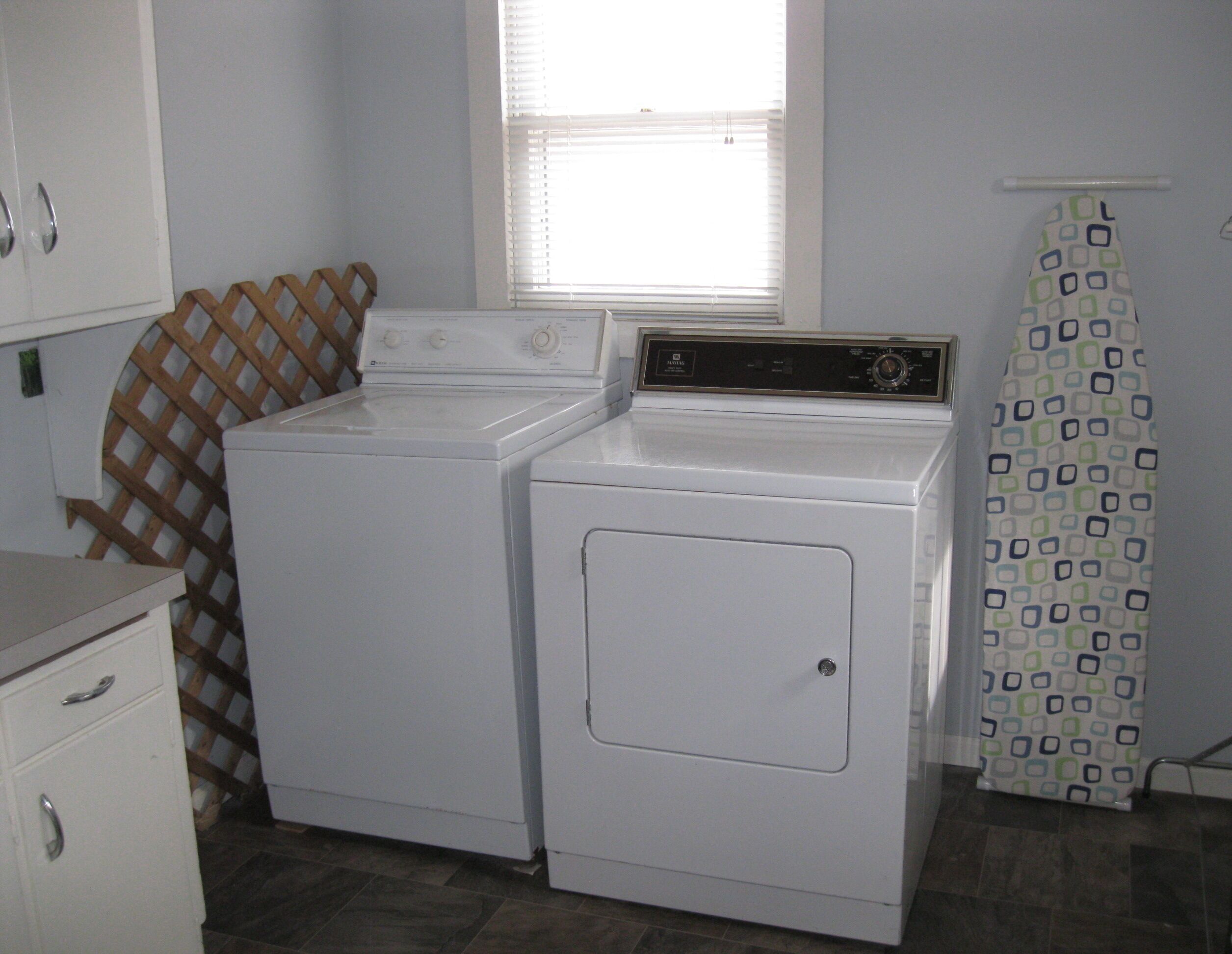 Une buanderie équipée de deux machines à laver et de deux sèche-linge superposés, d’un mur en treillis de bois, d’une fenêtre avec des stores et d’un rideau de douche à motifs.