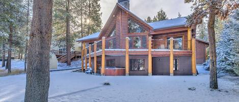 A log cabin with a large deck, surrounded by snow-covered trees.
