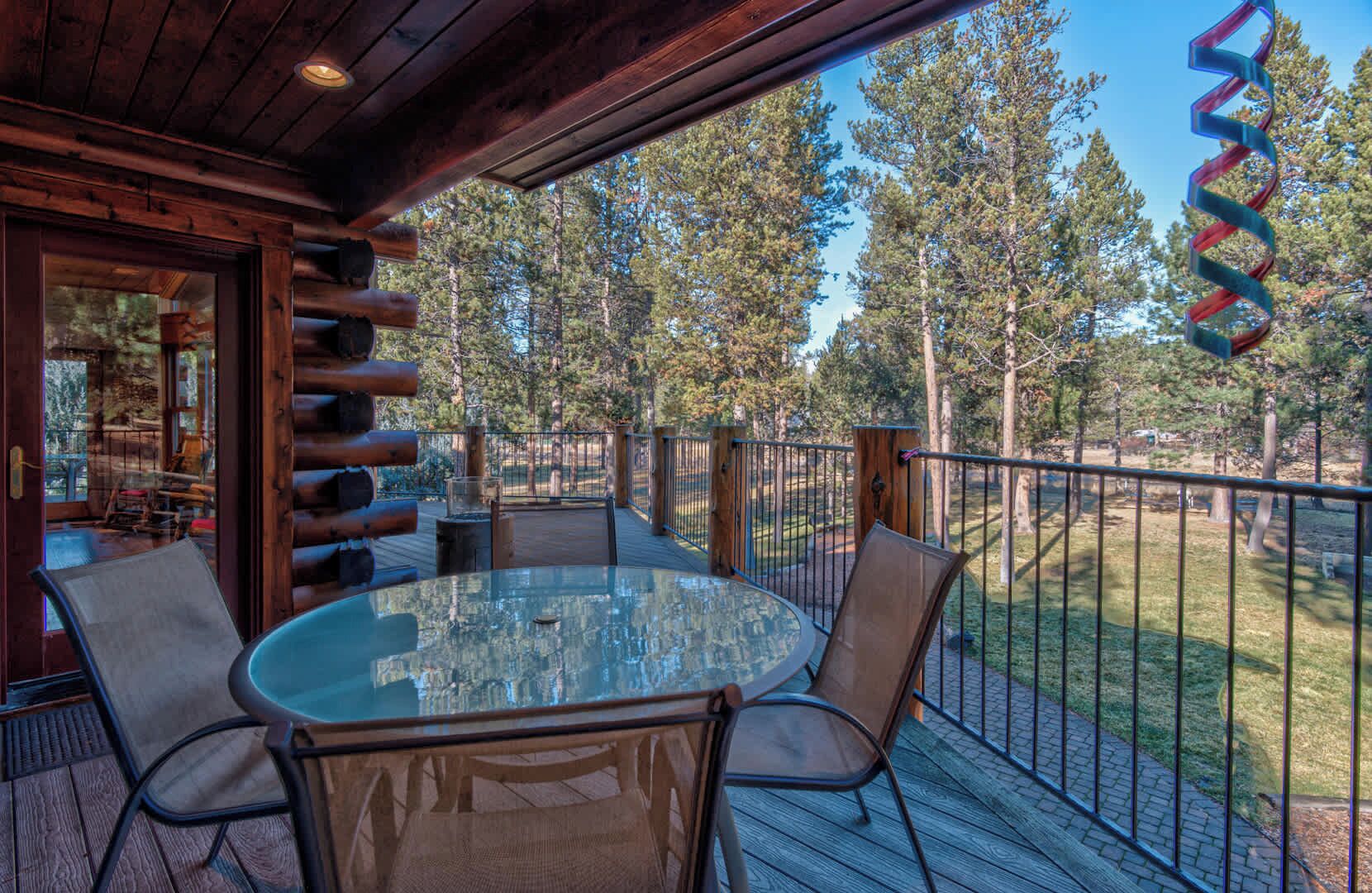 A wooden deck with a glass table and chairs, surrounded by a metal railing, overlooking a wooded area.