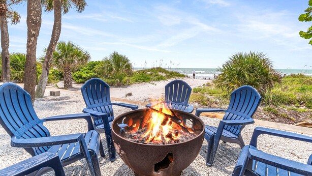 A beachside seating area with a fire pit, surrounded by chairs, palm trees, and a view of the ocean.