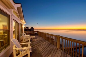 Une terrasse en bois avec des fauteuils Adirondack, une rambarde et une vue sur l’océan au coucher du soleil.