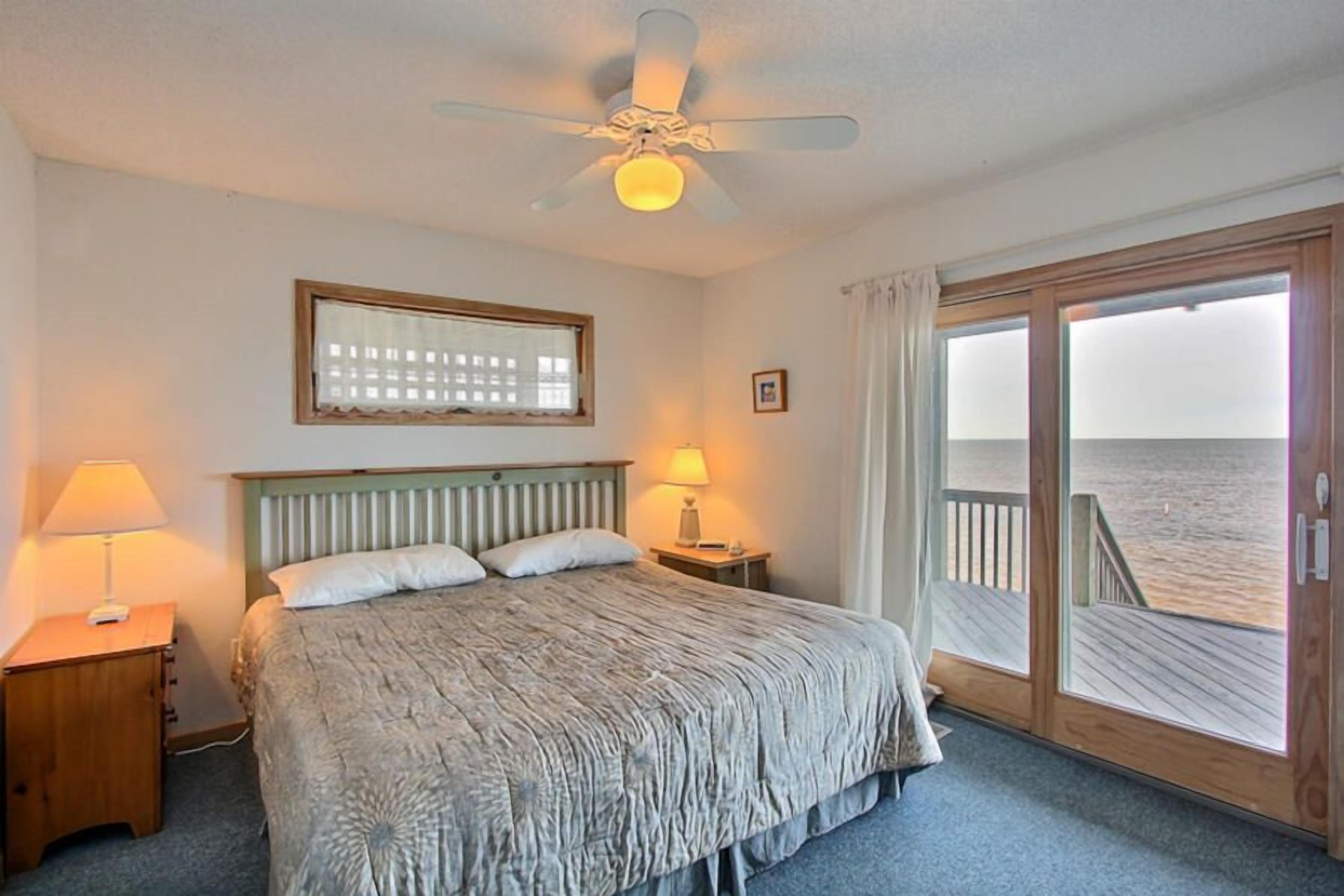 A bedroom with a bed, a ceiling fan, a window with curtains, a wooden deck, and a view of the ocean.