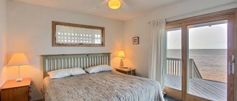 A bedroom with a bed, a ceiling fan, a window with curtains, a wooden deck, and a view of the ocean.