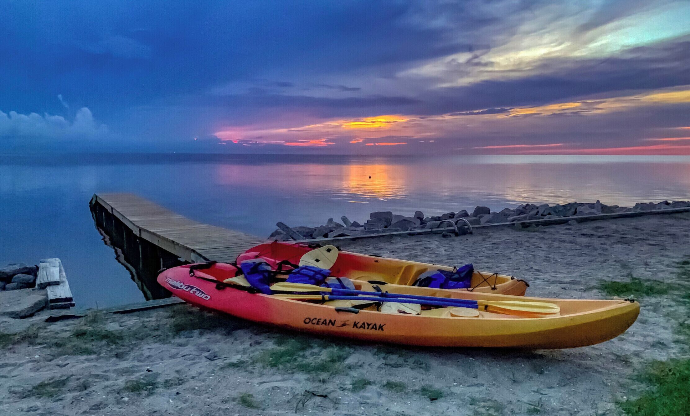 Een kajak met peddel en reddingsvest op een steiger aan een rustig water tijdens de zonsondergang.