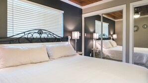 A bedroom with a large bed, two bedside tables with lamps, a mirror, and a window with blinds. - Modern 3 Bedroom Family Friendly Kihei Home Near Beach (Kihei)