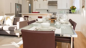 Una cocina moderna con armarios blancos, electrodomésticos de acero inoxidable y un comedor con mesa de vidrio y sillas marrones.