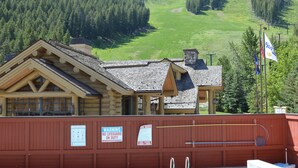 Area piscina con vista su una valle boscosa, un edificio in legno con un cartello che indica assenza di bagnino e un’asta con delle bandiere.