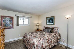 Een slaapkamer met een bloemenbedekking, een dressoir, een nachtkastje met een lamp, en twee ingelijste kunstwerken aan de muur.
