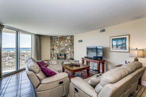 Een ruime woonkamer met een stenen openhaard, leren banken, een televisie en een groot raam met uitzicht op het strand.