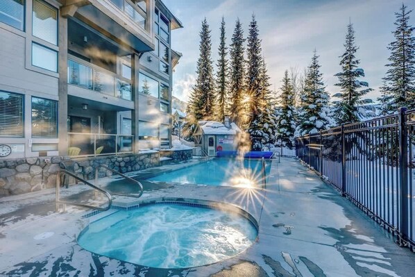 Ein Poolbereich mit Whirlpool, umgeben von einem Gebäude mit Balkonen und schneebedeckten Bäumen.