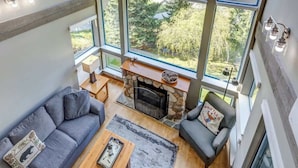 A spacious living room with a stone fireplace, large windows, and a view of a forest. - Greystone Lodge 311 Ski-in/Out by Acer Vacations (Whistler)