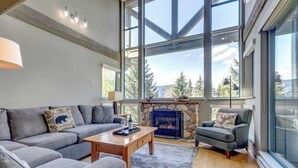 A modern living room with a stone fireplace, large windows, and a cozy seating area. - Greystone Lodge 311 Ski-in/Out by Acer Vacations (Whistler)