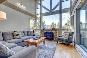 A modern living room with a stone fireplace, large windows, and a cozy seating area. - Greystone Lodge 311 Ski-in/Out by Acer Vacations (Whistler)