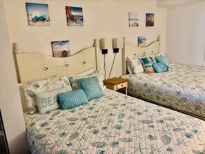 Twee eenpersoonsbedden met identiek bloemenbeddels, een nachtkastje en wanddecoratie met kusttaferelen.