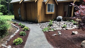 Una cabaña de madera con un sendero de grava que conduce a la entrada, rodeada de un jardín ajardinado con diversas plantas y rocas.