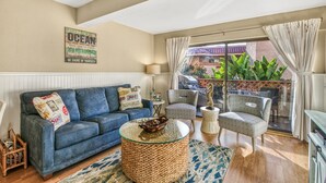 A living room with a blue sofa, a round coffee table, and two patterned chairs. A large window with curtains offers a view of greenery outside.