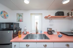 Een keuken met een roestvrijstalen spoelbak, een magnetron, een koelkast en een koffiezetapparaat. Er zijn witte keukenkastjes en een roze aanrecht.