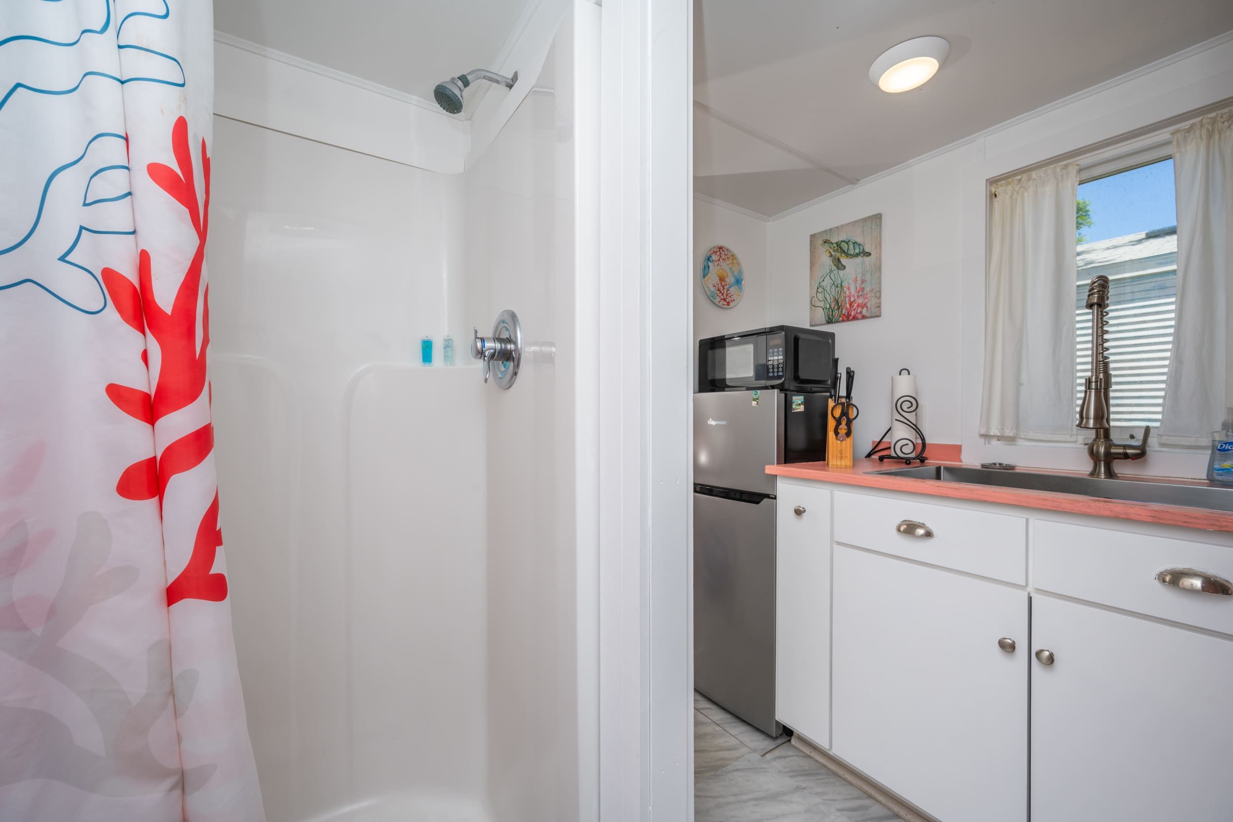 A bathroom with a shower and a toilet. There is a kitchen area with a microwave, sink, and cabinets.