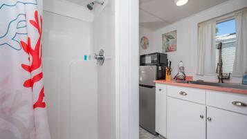 A bathroom with a shower and a toilet. There is a kitchen area with a microwave, sink, and cabinets.