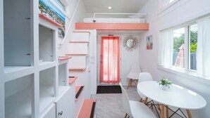 A compact hotel room with a bed, a small dining table, a chair, a white wall with a painting, a mirror, and a window with curtains.