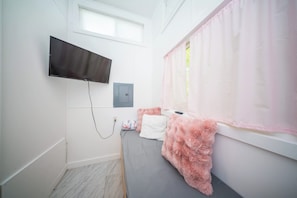 Een slaapkamer met een bed, een aan de muur bevestigde televisie, een raam met roze gordijnen en een stopcontact.