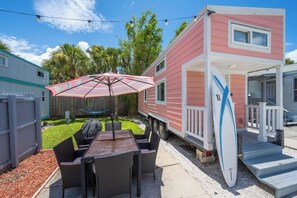 Een roze stacaravan met een patio, inclusief een tafel, stoelen, een gestreept zonnescherm en een surfboard tegen de caravan geleund.