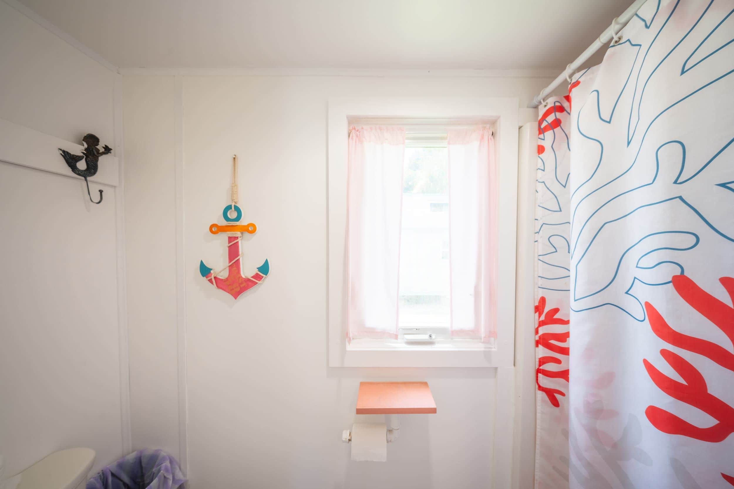 Ein Badezimmer mit Fenster, einer Duschvorhang mit Blättermuster, einem Toilettenpapierhalter und einem kleinen Regal.