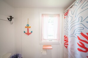 Un baño con ventana, cortina de ducha estampada con hojas, dispensador de papel higiénico y un estante pequeño.