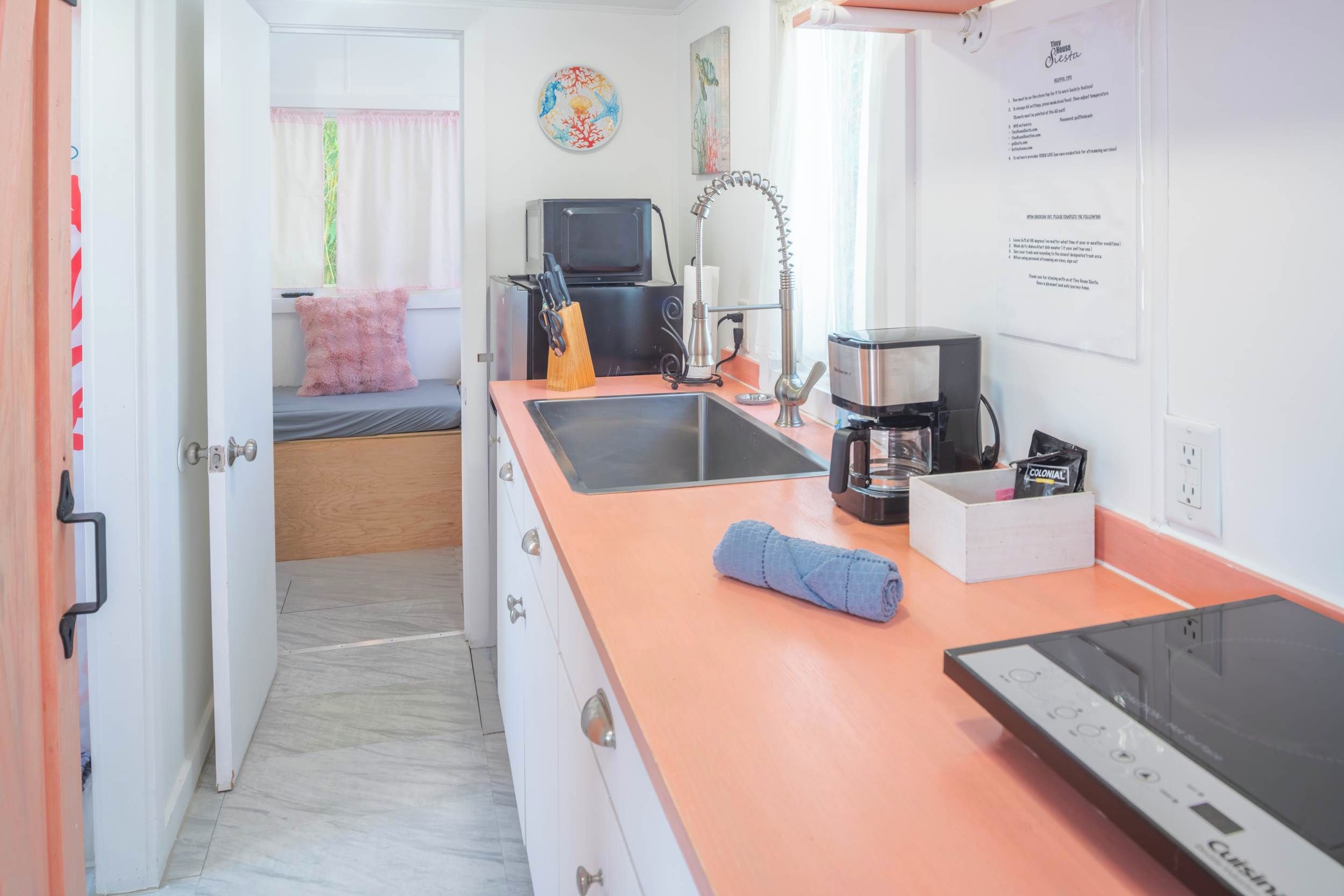 Eine kleine Küche mit Arbeitsplatte, Spüle und Geräten. Im Hintergrund ist ein Bett mit einem rosafarbenen Kissen zu sehen.